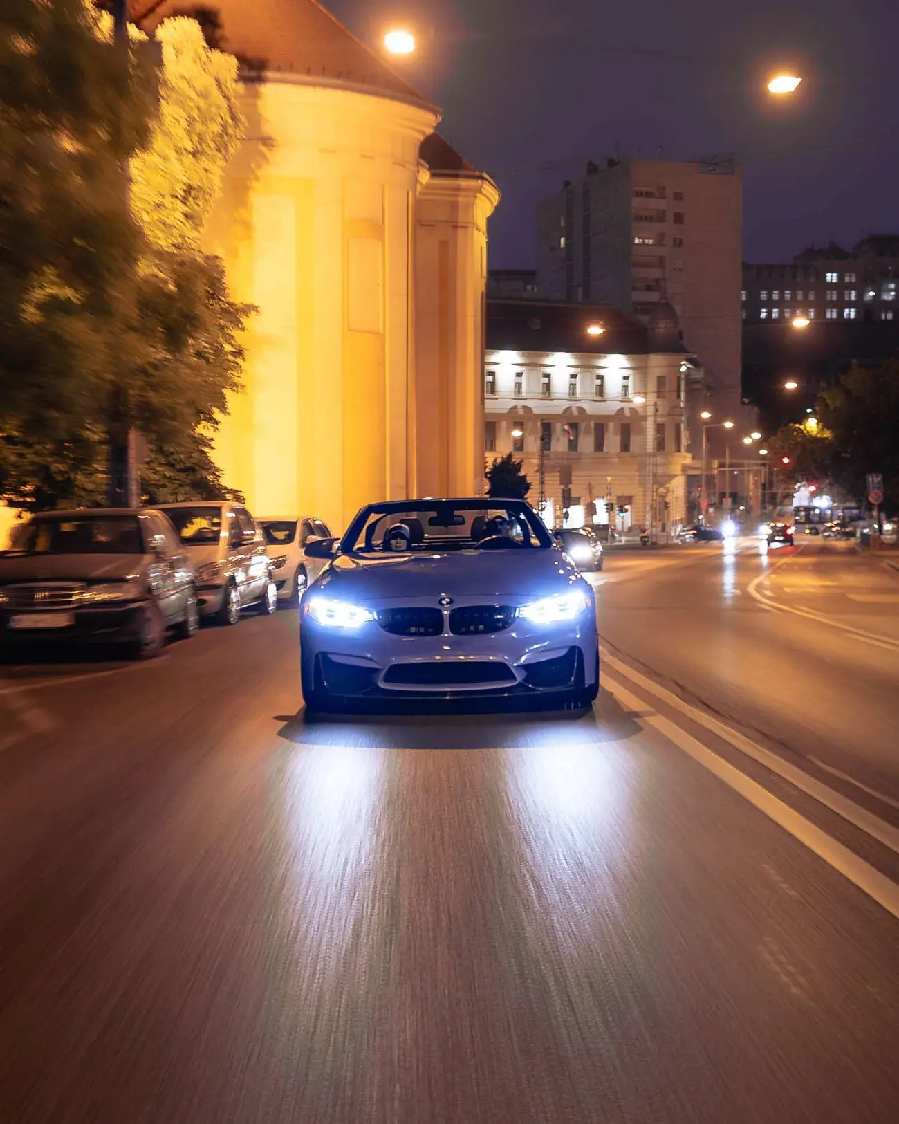 Blue BMW M4 Convertible at night, headlights on, a floodlit building behind