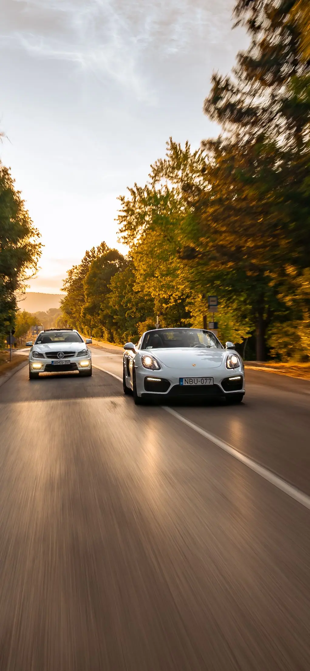 Mercedes-AMG C 63 and Porsche Boxster GTS side by side on a tree-lined road at sunset
