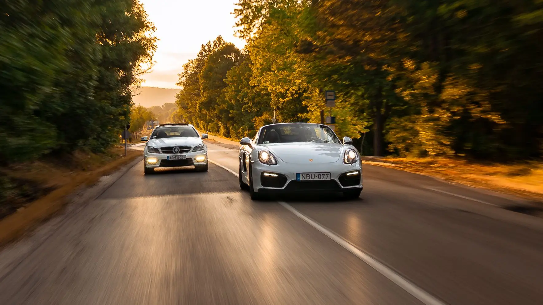 Mercedes-AMG C 63 and Porsche Boxster GTS side by side on a tree-lined road at sunset