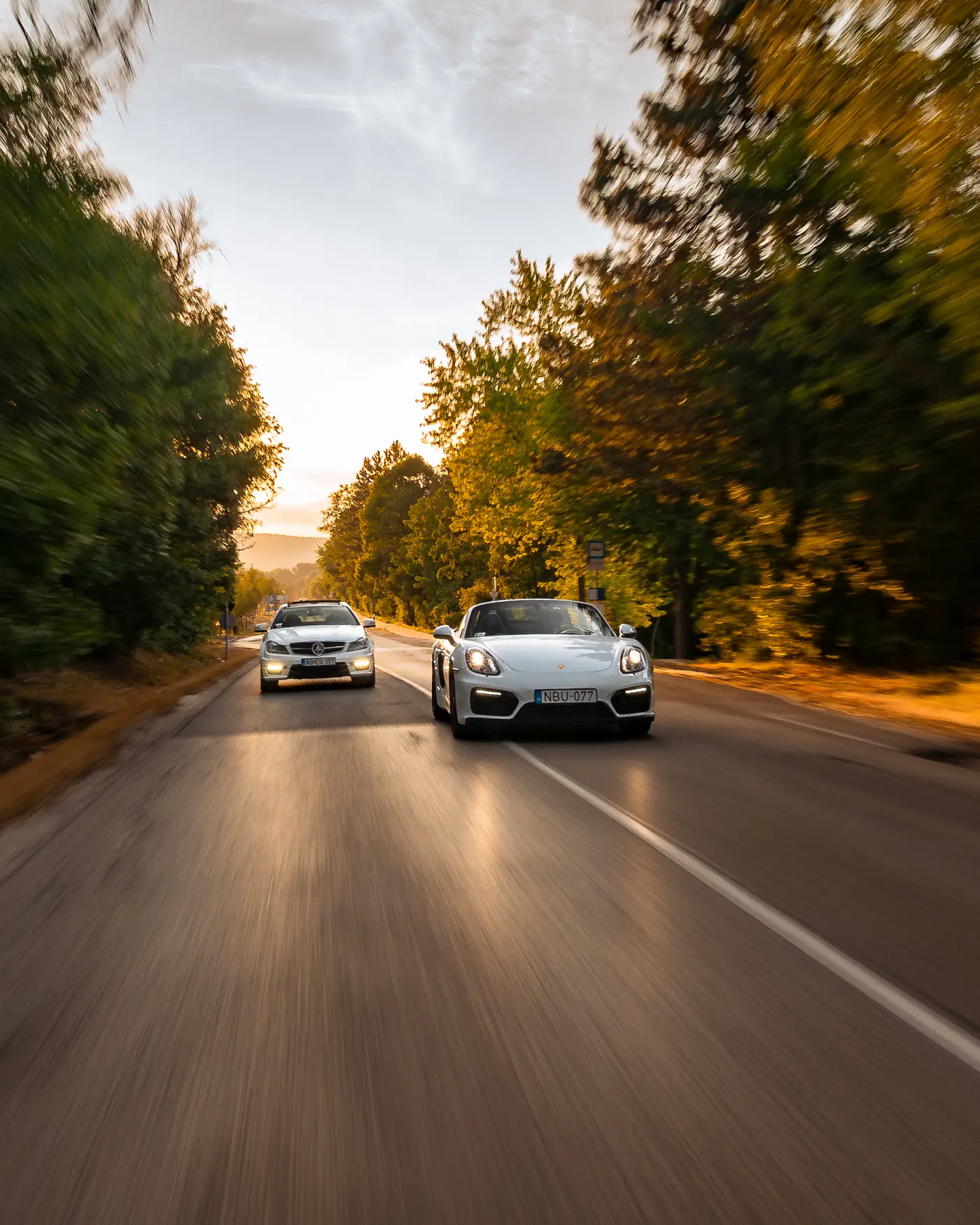 Mercedes-AMG C 63 and Porsche Boxster GTS side by side on a tree-lined road at sunset