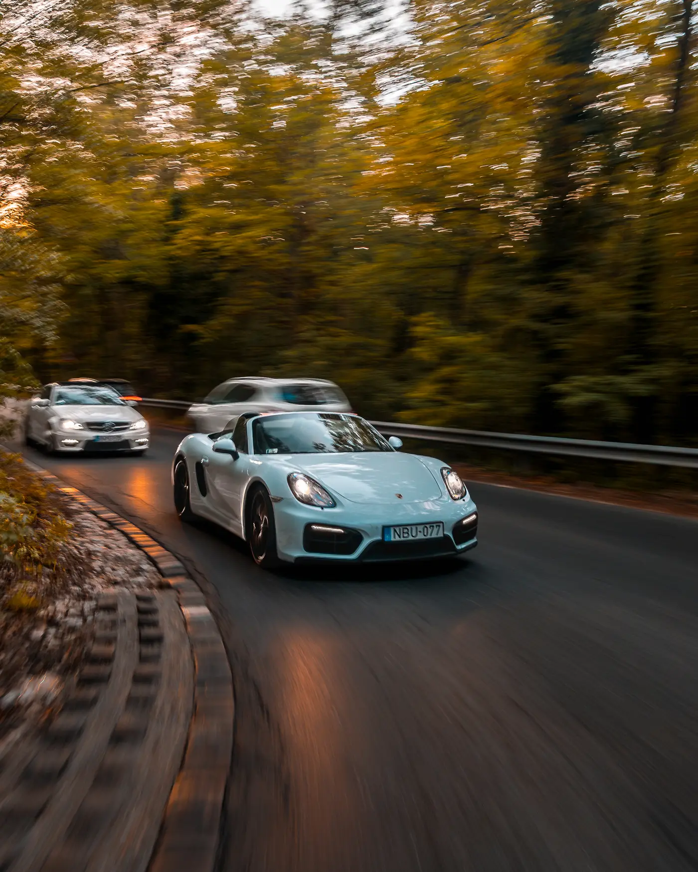 Porsche Boxster GTS leading the Mercedes-AMG C 63 through a bend, guardrail and blurred trees behind