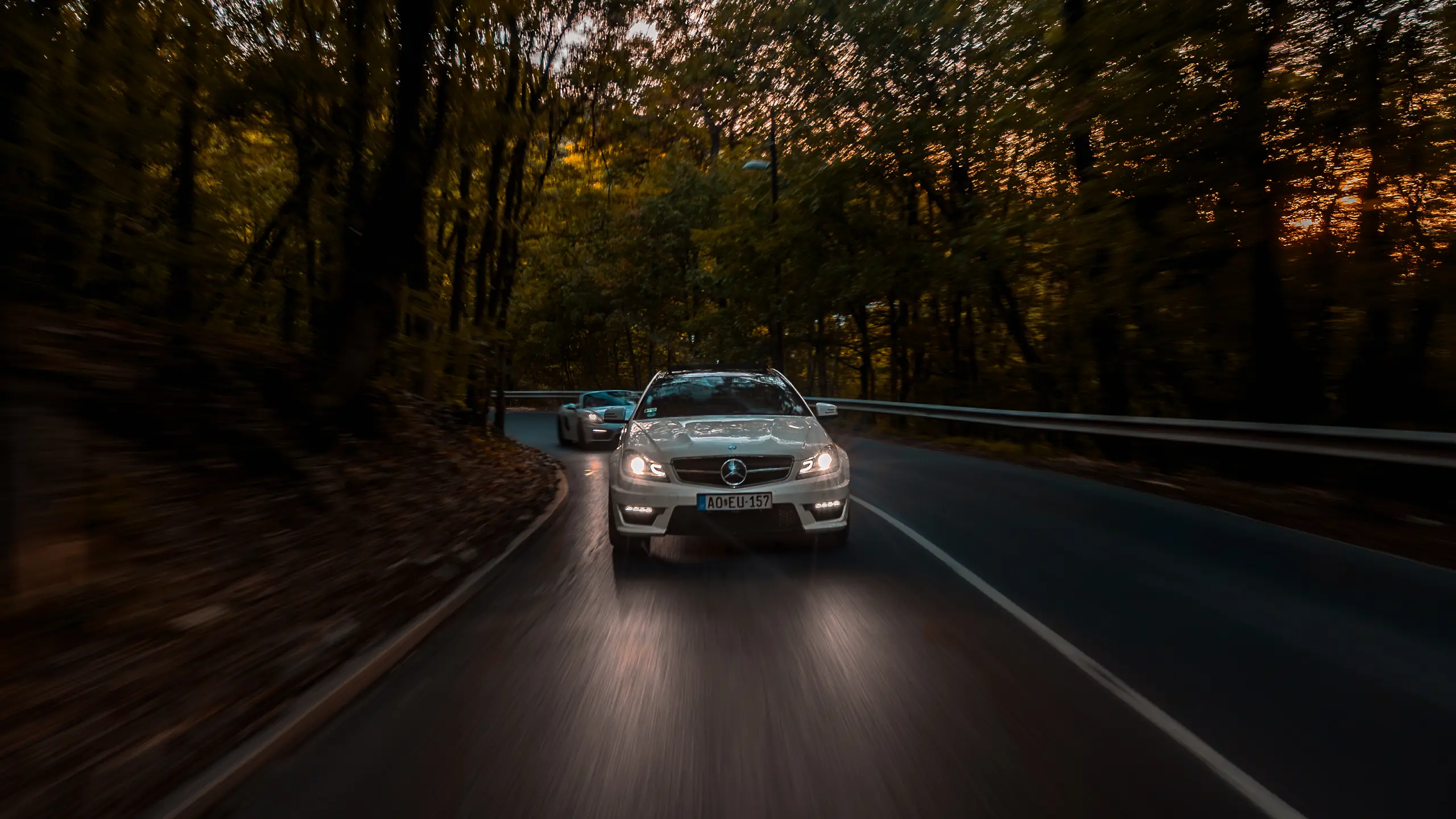 Mercedes-AMG C 63 leading a Porsche Boxster GTS on a forest road at dusk