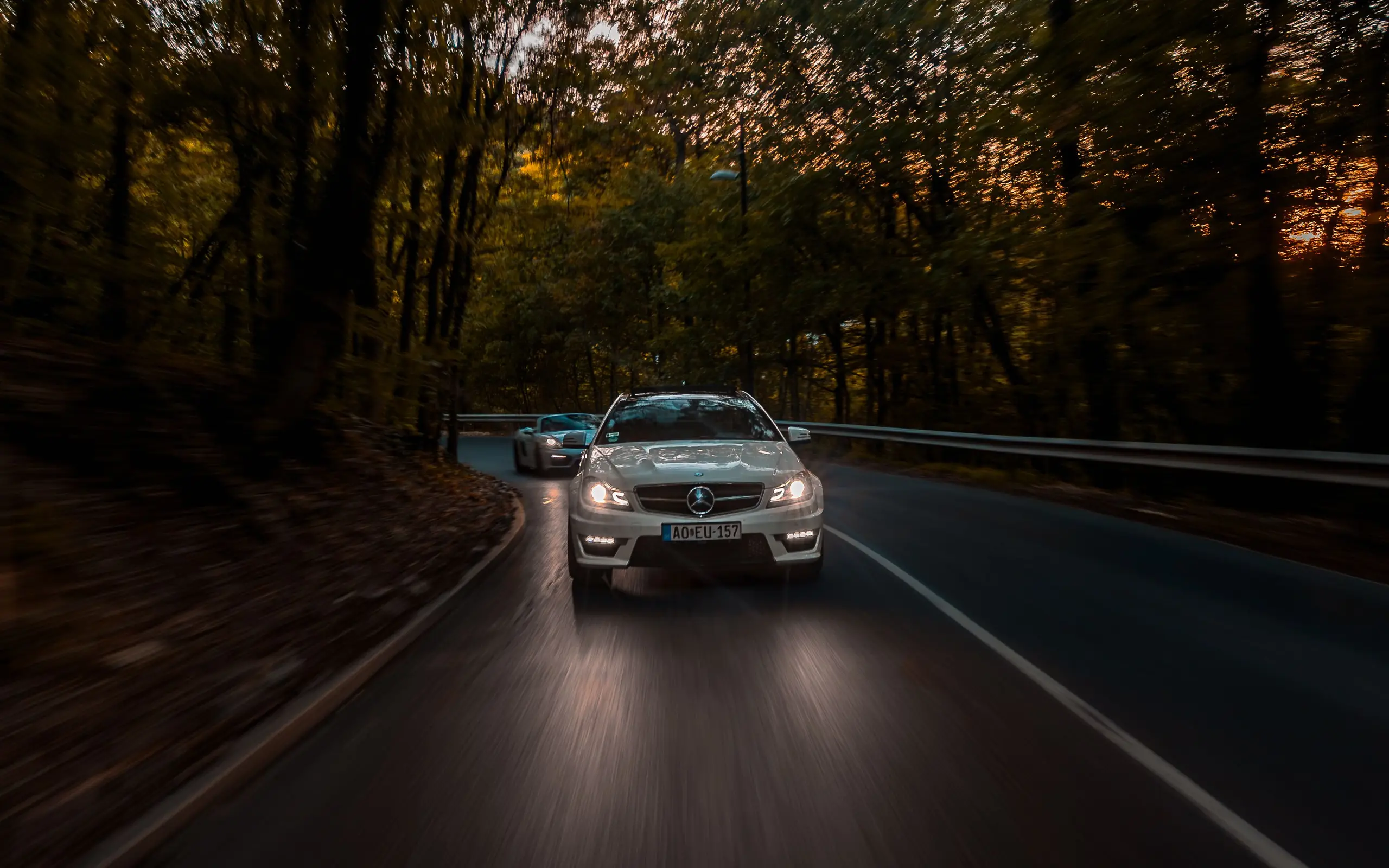 Mercedes-AMG C 63 leading a Porsche Boxster GTS on a forest road at dusk