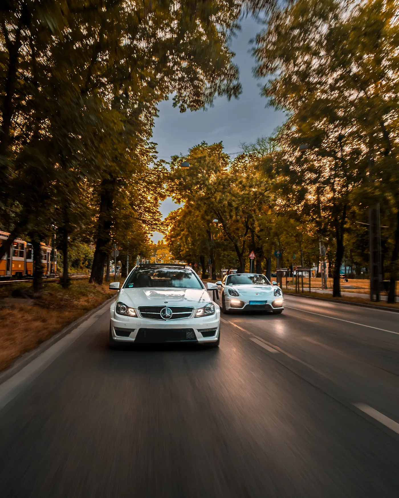 Mercedes-AMG C 63 and Porsche Boxster GTS on a tree-lined avenue, a tram behind