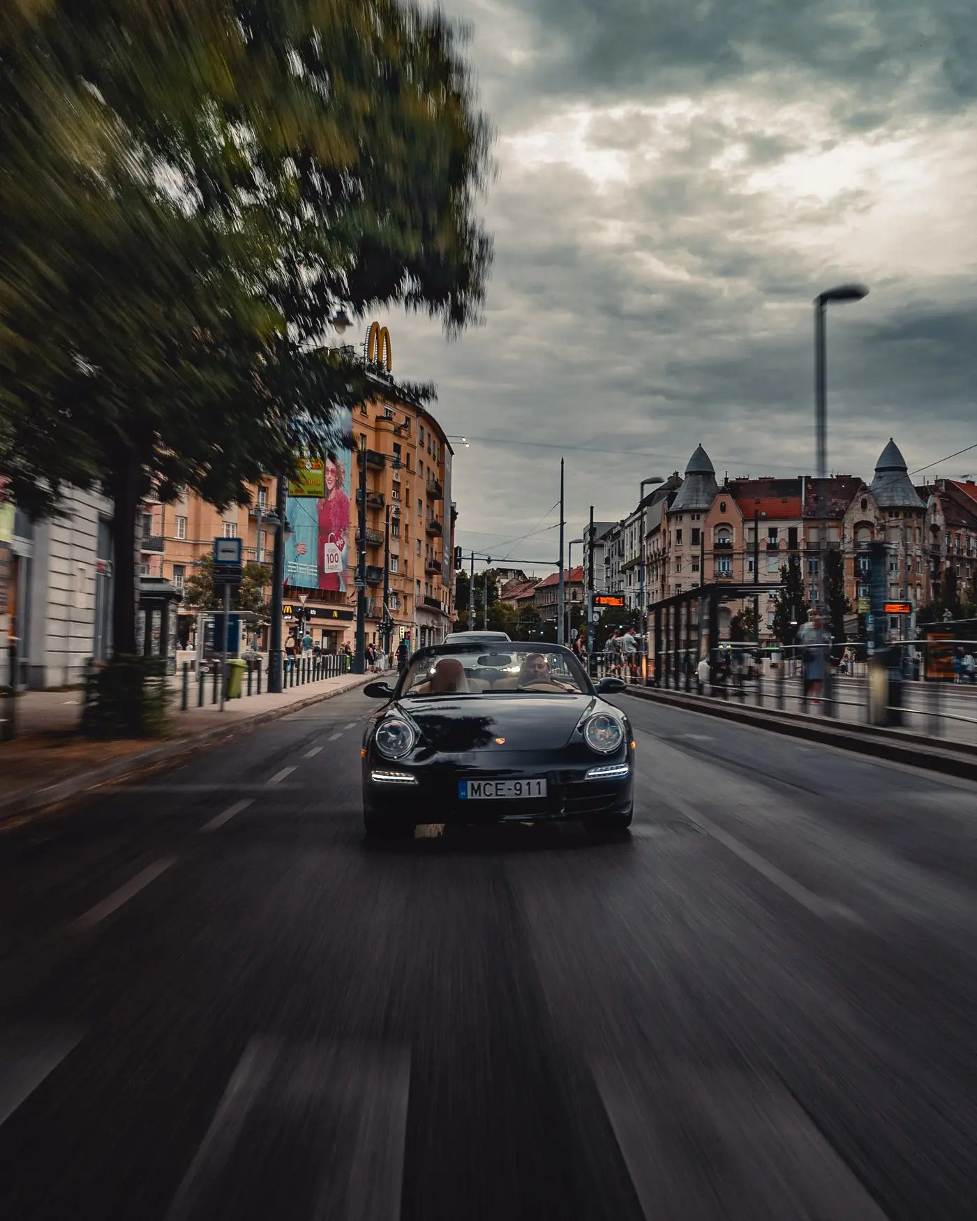 Porsche 911 Carrera Cabriolet head-on down a city avenue, trees blurred on the left