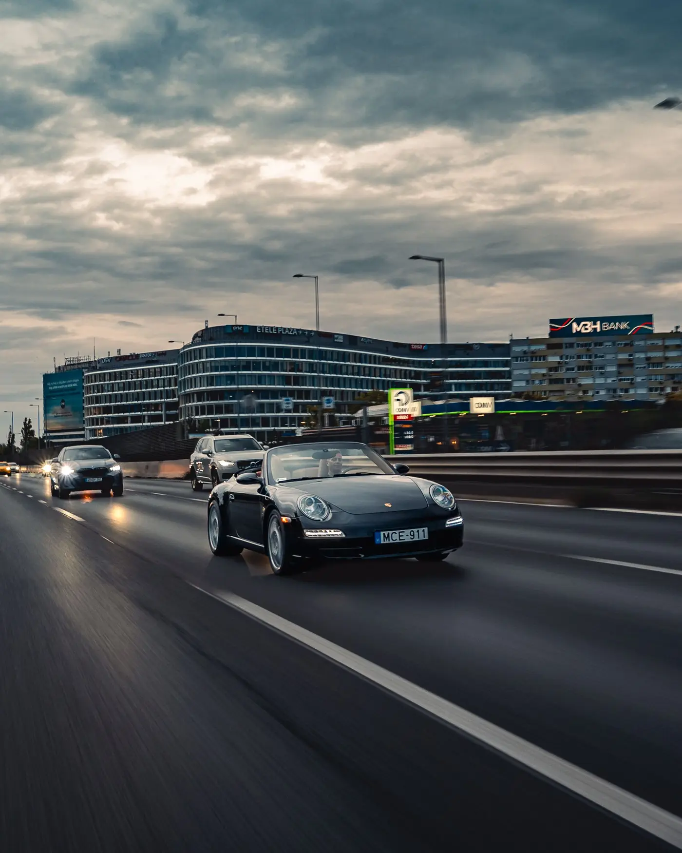 Porsche 911 Carrera Cabriolet on a multi-lane road, office buildings and evening clouds behind
