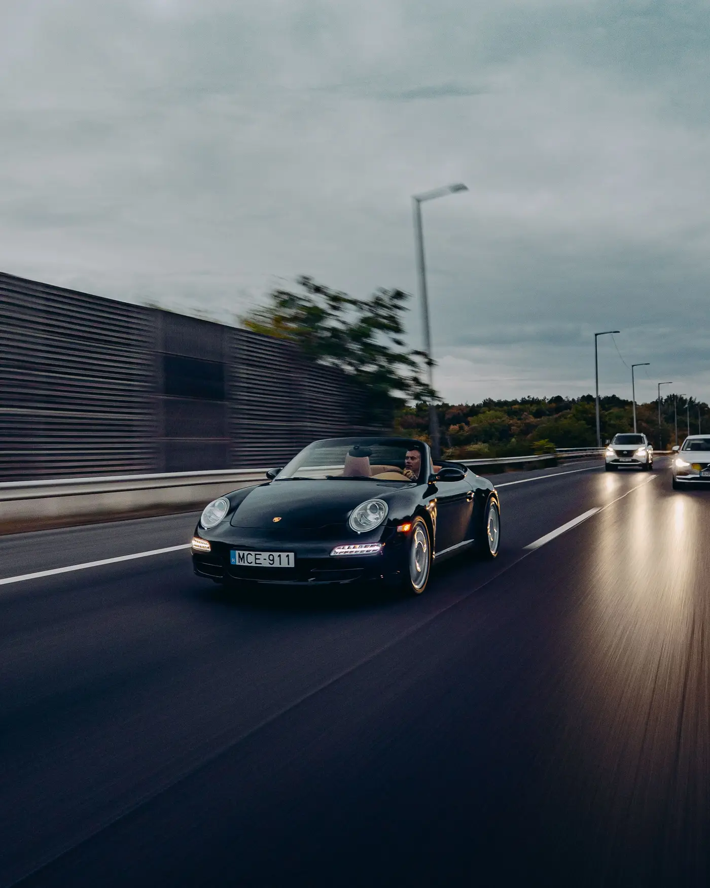 Porsche 911 Carrera Cabriolet on a motorway beside a noise barrier, headlights following