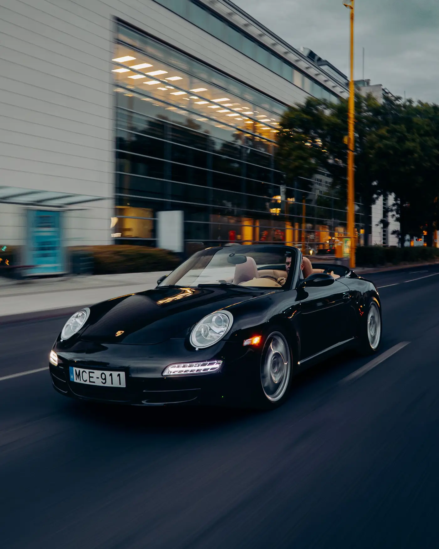 Porsche 911 Carrera Cabriolet passing a lit glass building at dusk