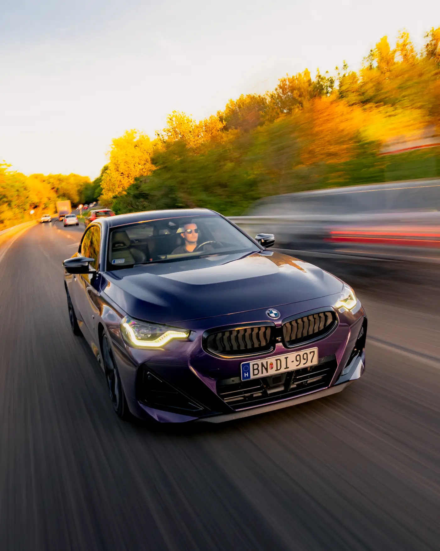Purple BMW M240i in traffic on a road lined with autumn trees