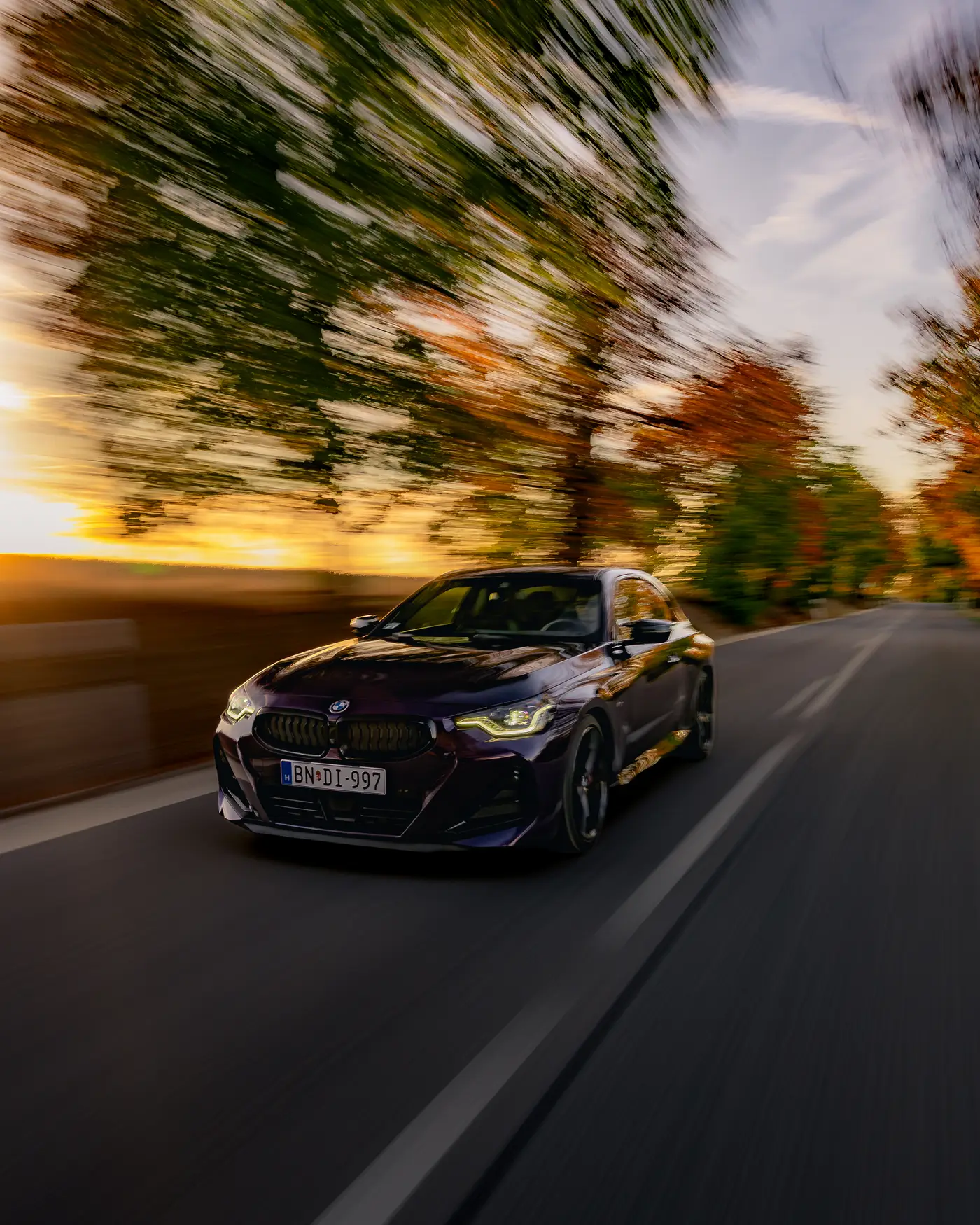 BMW M240i under trees blurred by motion, low sun behind