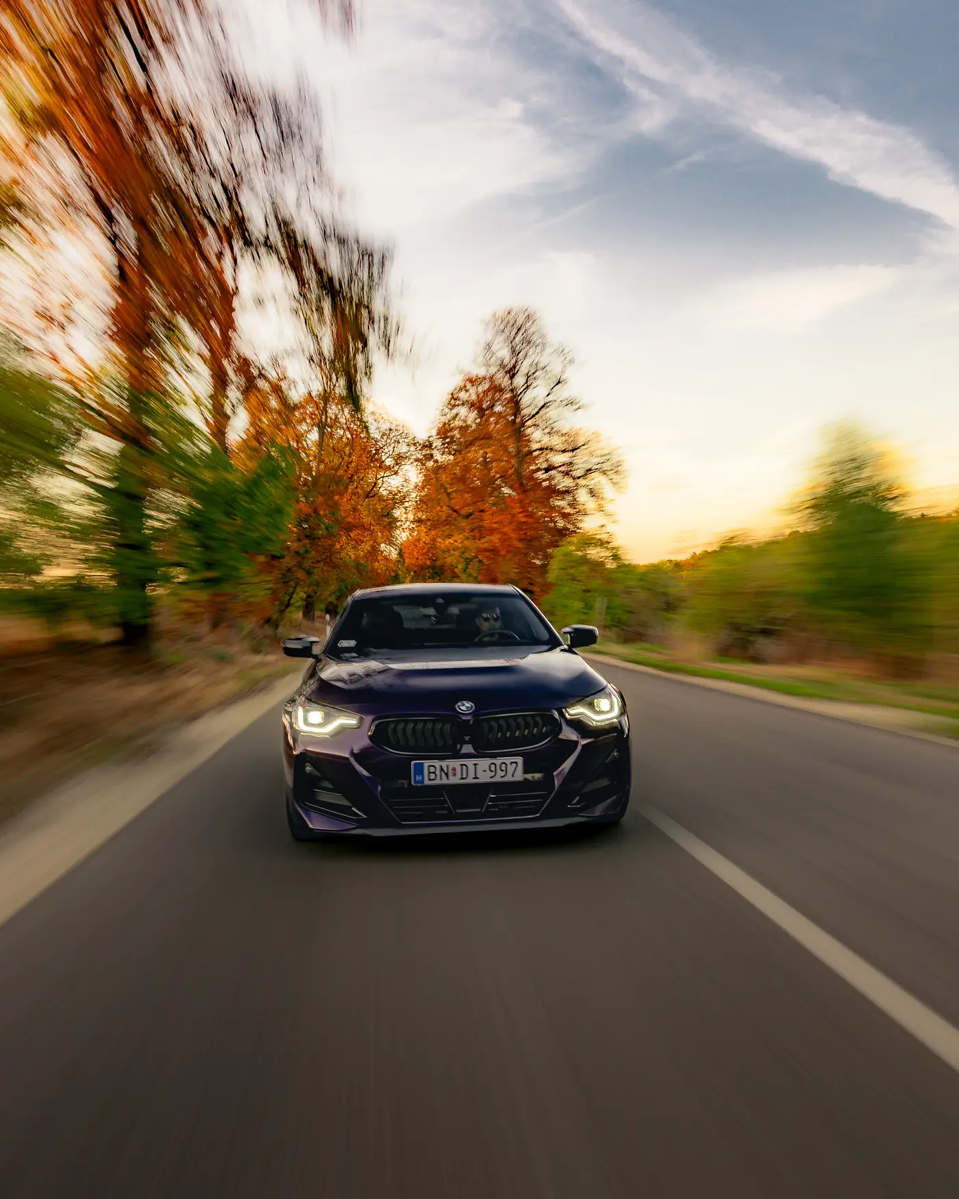 BMW M240i under autumn trees blurred by motion, evening sky