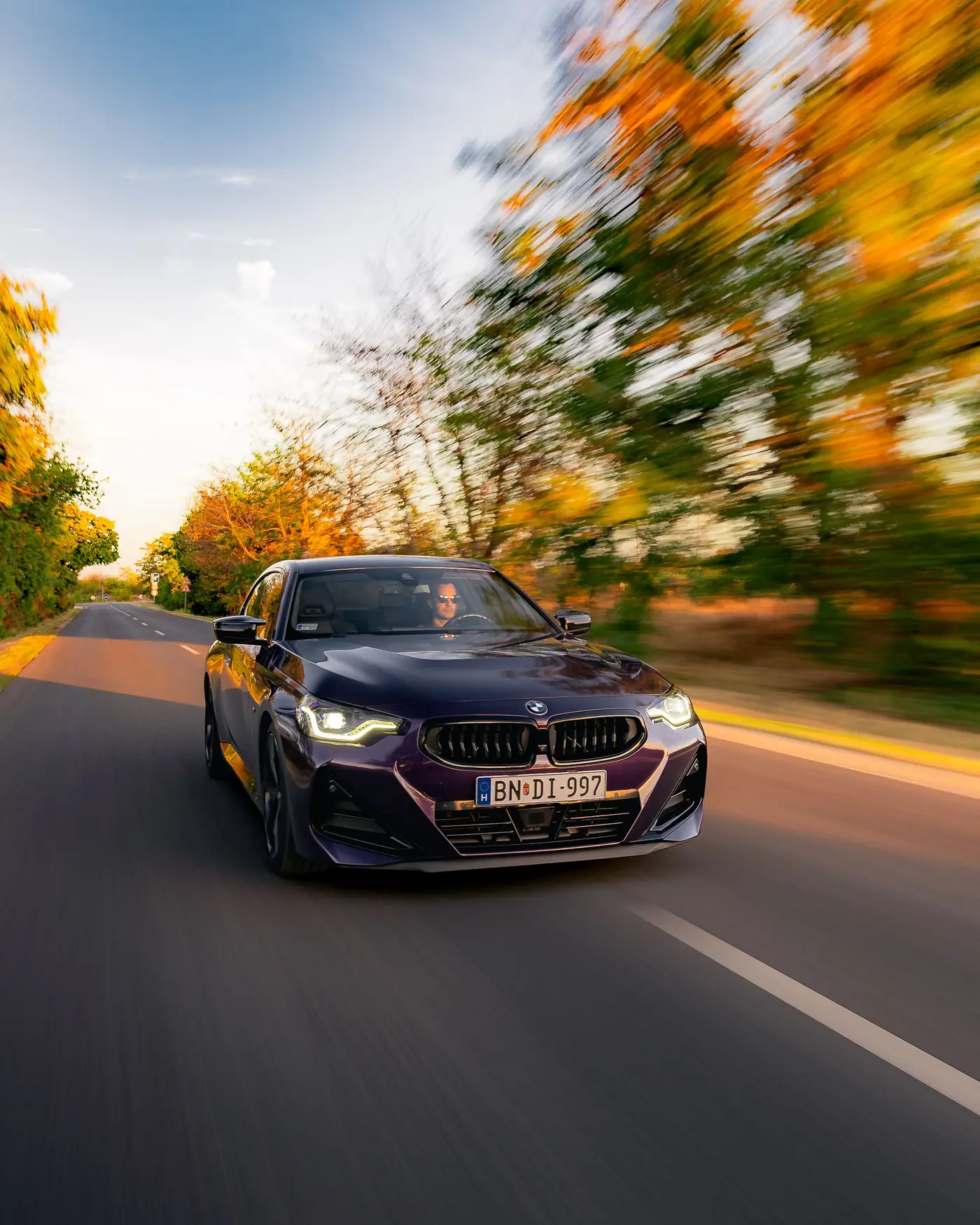 BMW M240i close up, green and orange trees blurred against a blue sky