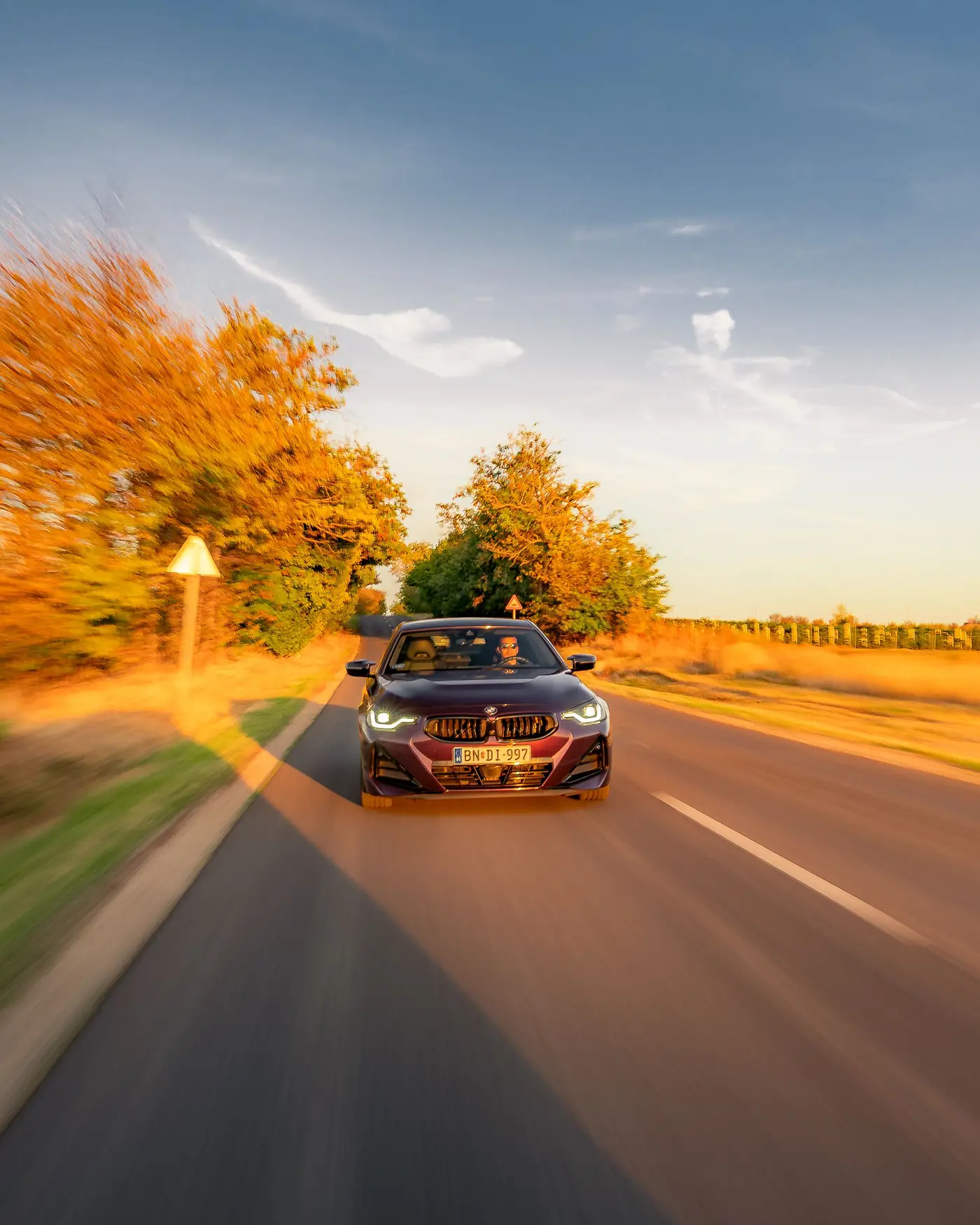 BMW M240i in low sun on a country road, a road sign and autumn trees beside it